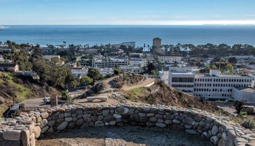 View from a hilltop showing a coastal cityscape with buildings, roads, and a pier extending into the ocean under a clear blue sky. The ideal location to plan your event, with a rock wall in the foreground adding rustic charm.