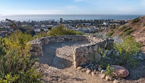 A stone-paved lookout point with circular seating on a hill overlooks a coastal town and the ocean in the distance, providing a perfect spot to plan your next event on a clear day.