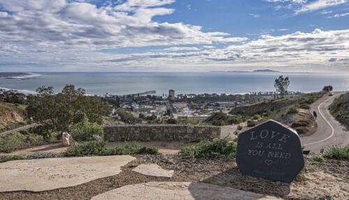 Scenic view of a coastal town with an ocean in the background, taken from a hilltop. A stone inscribed with "LOVE IS ALL YOU NEED" is visible in the bottom right corner of the foreground, ideal for planning your event against a breathtaking backdrop.