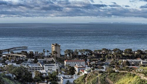 Coastal cityscape view with buildings, roads, and greenery in the foreground, overlooking a calm ocean and an overcast sky. A pier extends into the water on the left. Islands are visible in the distance, creating the perfect backdrop to plan your next event by the sea.