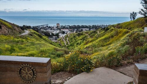 View of a coastal city from a hill with a Rotary Club plaque in the foreground, green hillsides, and a clear blue sky above the ocean. The serene setting makes it an ideal spot to plan your next event.