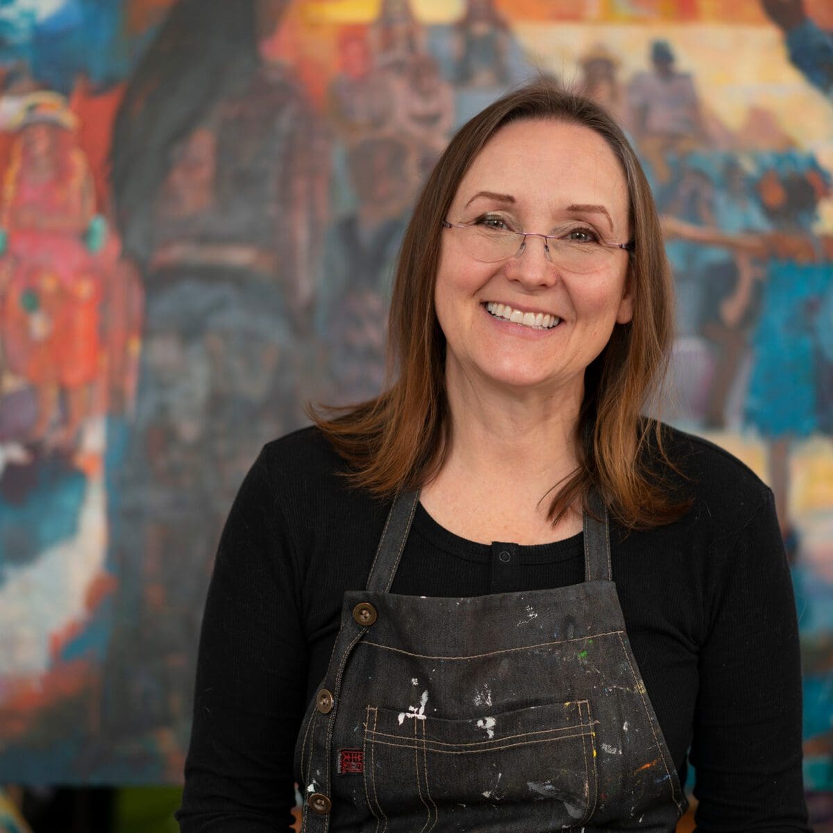 Anette Power, wearing glasses and a paint-stained apron, smiles in front of a colorful, abstract painting.