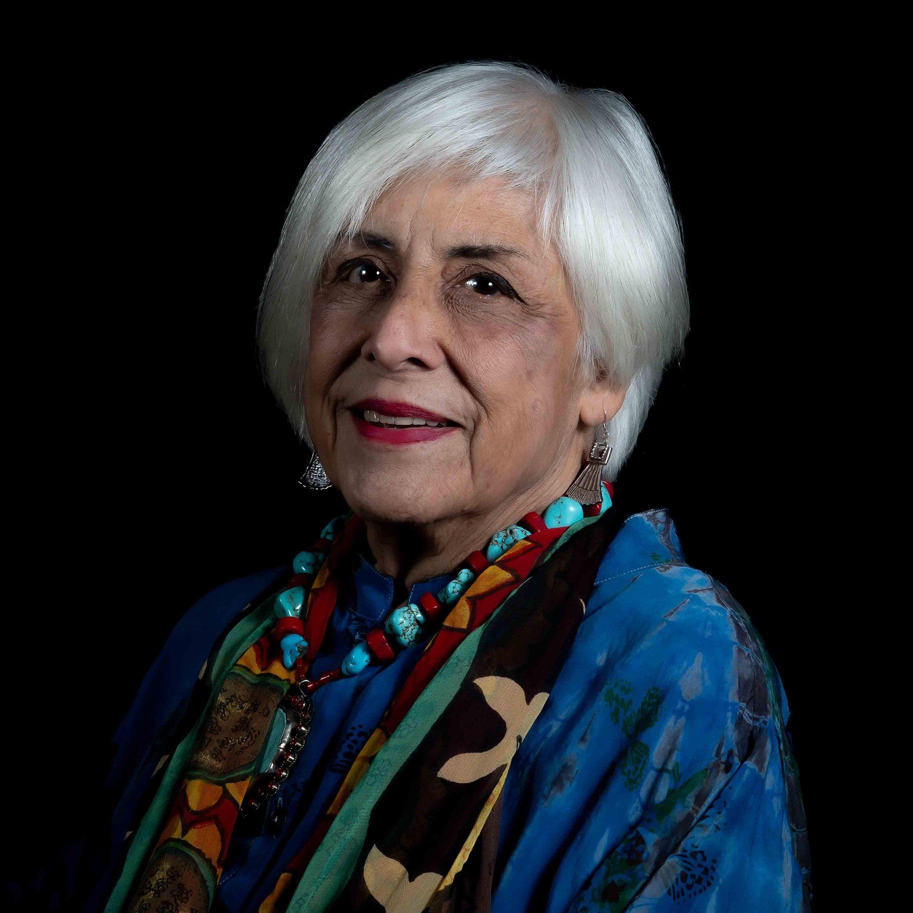 Margaret Garcia, an elderly woman with short white hair, wearing a blue top, a multi-colored scarf, and jewelry, poses gracefully against a dark background.