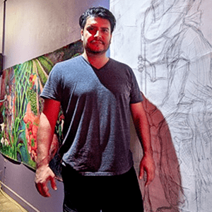 A man with dark hair and a beard, presumably Ruben Zavala, stands in front of vibrant and monochrome artworks on the wall. He is wearing a grey t-shirt and black pants.