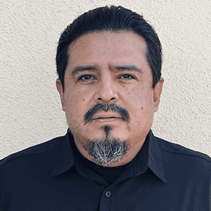 Jorge Beltran, a man with a goatee and mustache, stands against a light-colored wall dressed in a black shirt, looking directly at the camera.