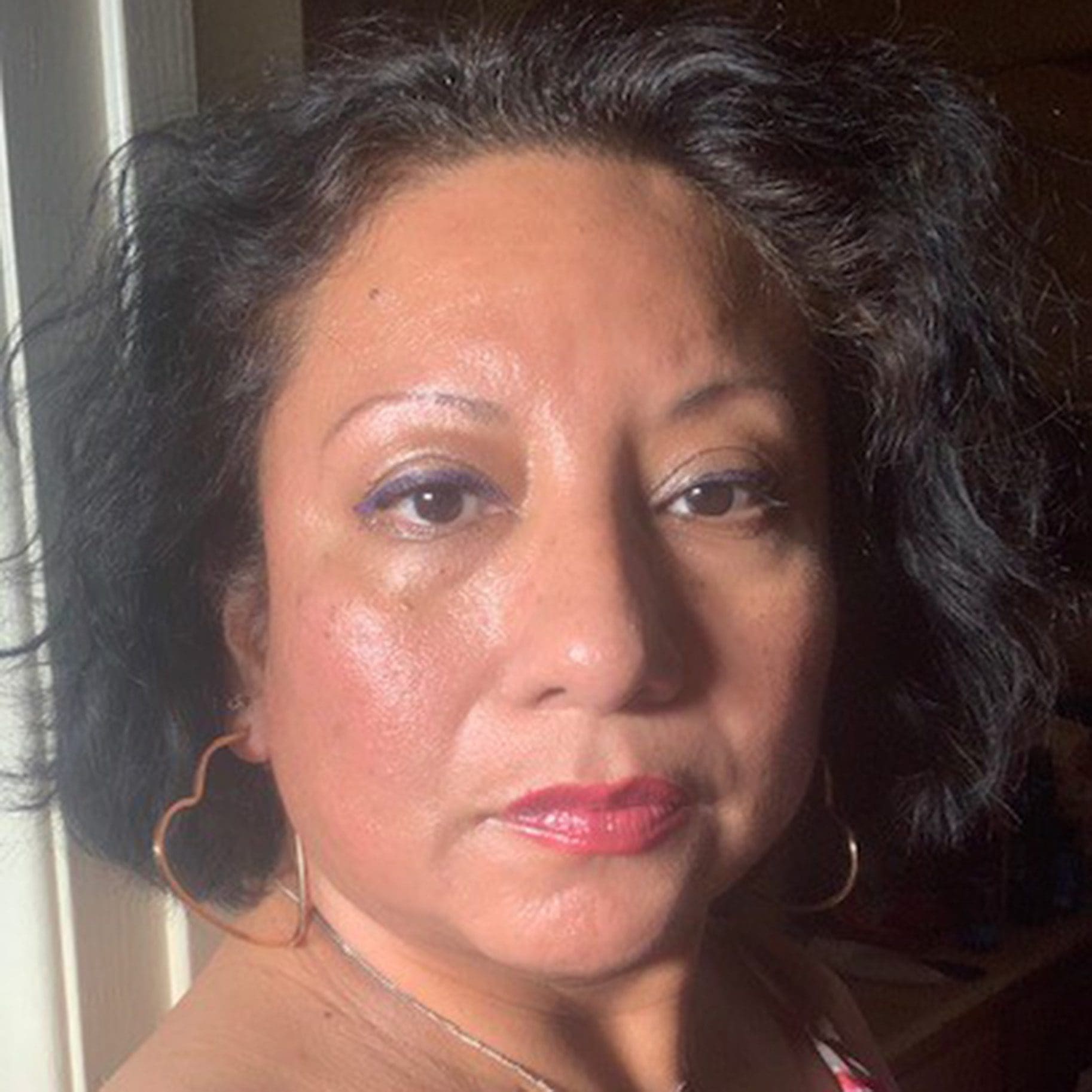 A woman with short dark hair, wearing large heart-shaped earrings and a pink top, looks directly at the camera under bright lighting. Gloria Vasquez-Warner's confident gaze draws viewers in effortlessly.