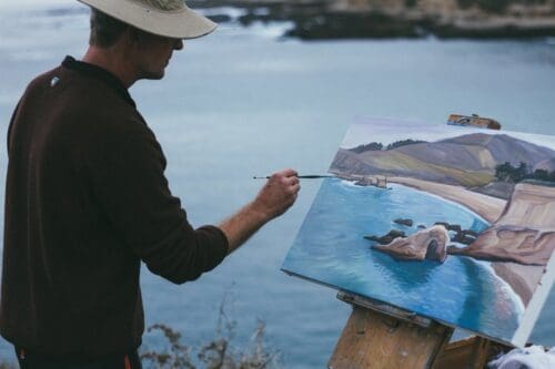 Jeremy Harper, in a hat and dark sweater, paints a coastal landscape on an easel next to a body of water.