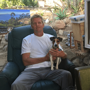 Jeremy Harper, a man in a white T-shirt and gray pants, sits in a dark green chair outdoors, holding a small dog.