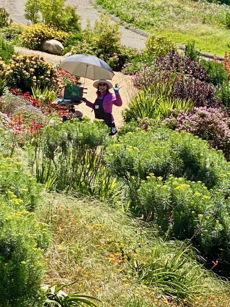 A person in a purple shirt and wide-brimmed hat stands in a garden, holding an umbrella and waving, surrounded by various plants and flowers, exuding an aura of creativity like an artist bringing a canvas to life.