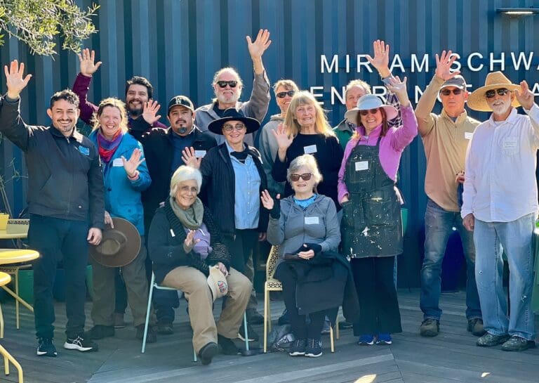 A group of 14 people, some standing and some sitting, smiling and waving at the camera. They are gathered in front of a building with signage reading "Miriam Schwab Center...". Among them are artists showcasing their creative spirit.