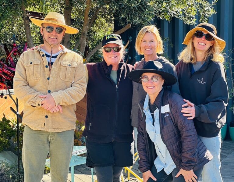 A group of five people, three women and two men, stand outdoors smiling. They are dressed casually with some wearing hats and sunglasses. Among these artists, trees and plants are visible in the background.