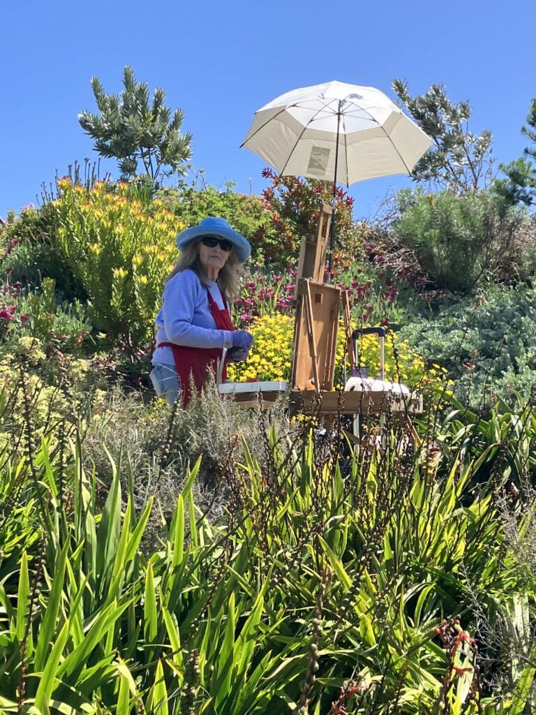 A creative artist wearing a blue hat and red apron paints at an easel under a white umbrella in a sunny, lush garden.