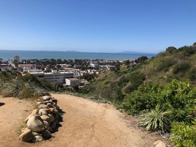 A sandy path with rocks on the left leads down a hillside covered in greenery, overlooking a coastal town and the ocean. With islands visible in the distance under a clear blue sky, it’s an inspiring scene that has captivated many artists.