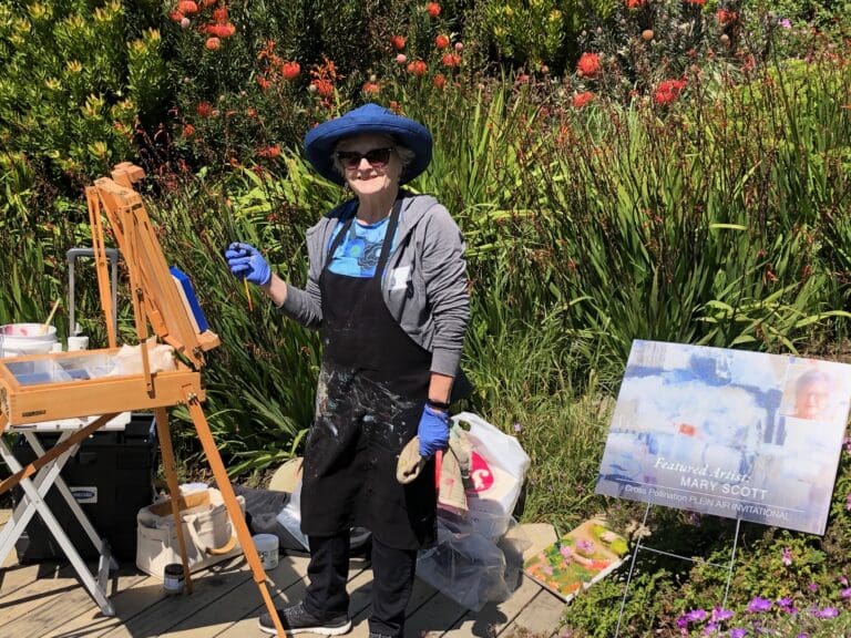 A person wearing a hat and apron stands painting at an easel outdoors, surrounded by art supplies and greenery. A sign next to them displays the name "Mary Scott," indicating the creative artist is a featured talent.