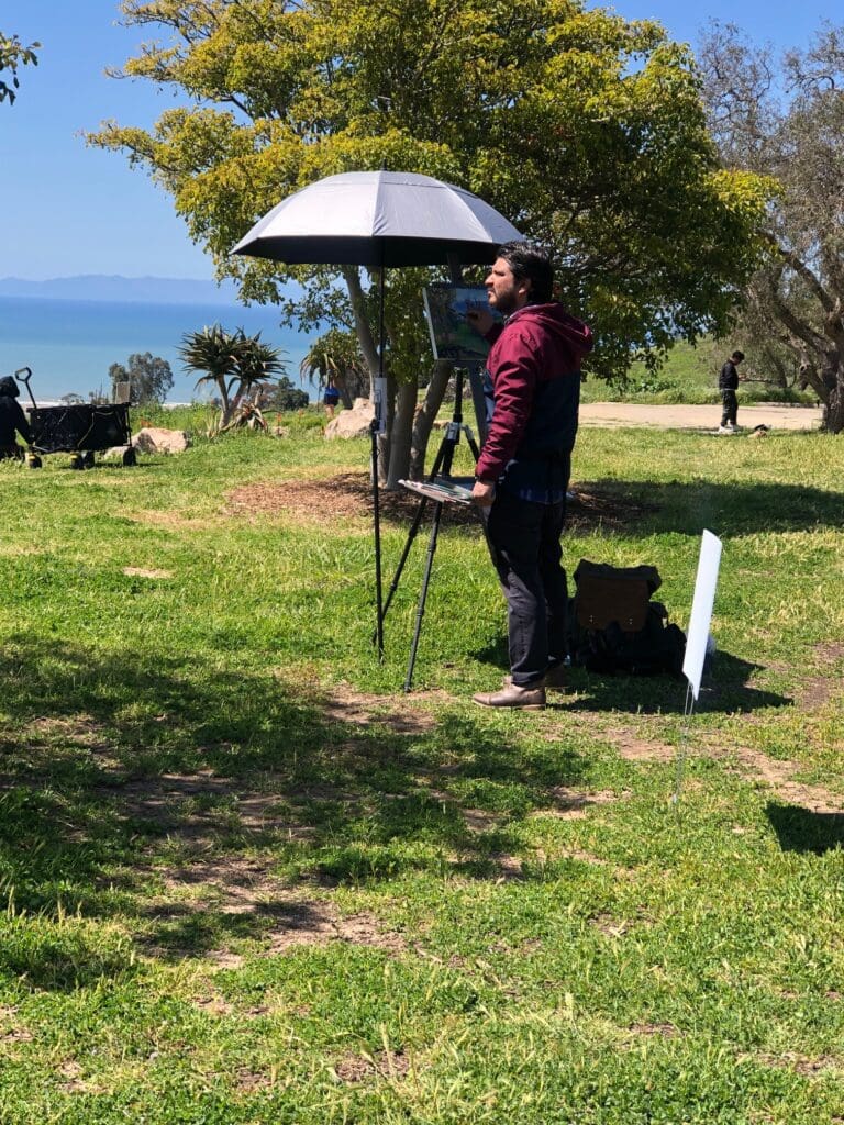 An artist is painting on an easel outdoors under an umbrella shade, with vibrant green grass, trees, and a distant view of the sea in the background.