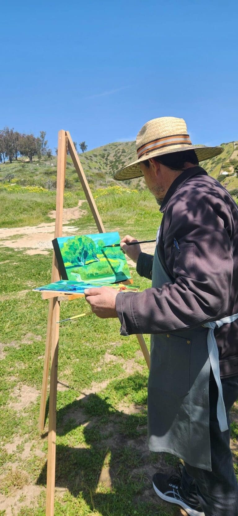 An artist wearing a hat and apron paints a landscape on an easel in an outdoor setting with green grass and hills in the background.