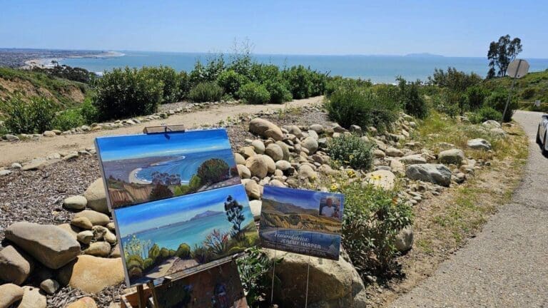 A scenic coastal view with an easel displaying two landscape paintings by local artists, one of which features the same coastal scene. Rocky terrain and vegetation are in the foreground, with the ocean and sky in the background.