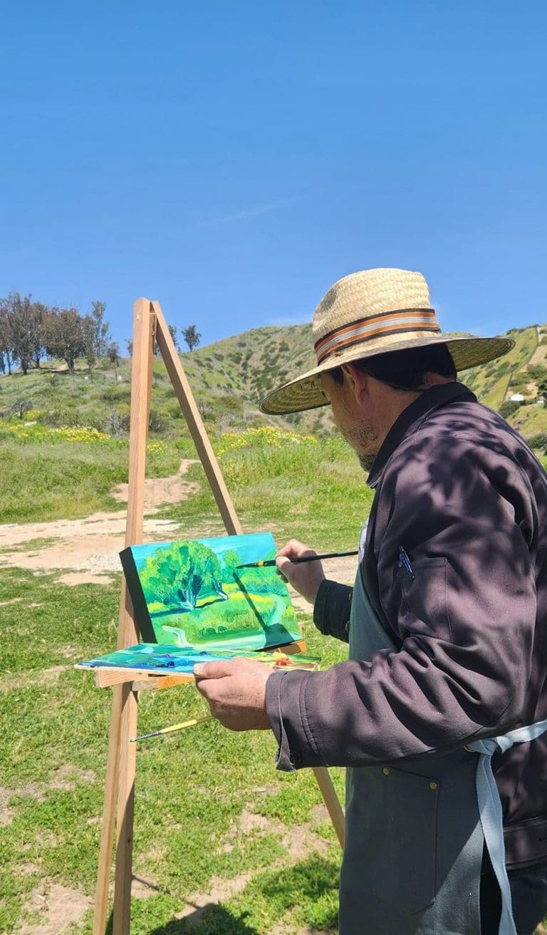 A person wearing a straw hat paints a landscape on an easel outdoors, under a clear blue sky with grassy hills and trees in the background.