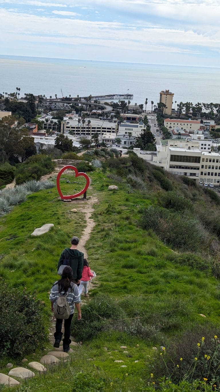 Participants in the In Bloom Contest stroll down a grassy hill, where a striking heart sculpture stands proudly. The backdrop of towering buildings and the vast ocean completes this picturesque scene.