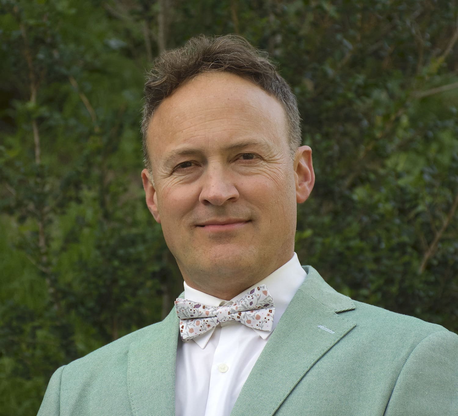 Man wearing a light green suit jacket, white shirt, and patterned bow tie, standing outdoors in front of green foliage.