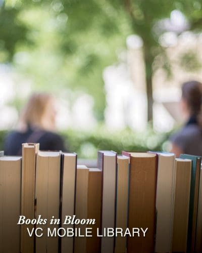 Several books stand upright in the foreground, with two people out of focus behind them. The image features the text: Books in Bloom VC MOBILE LIBRARY, highlighting the Books in Bloom community initiative.